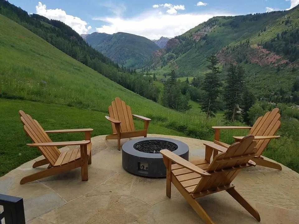 Four wooden Adirondack chairs around a fire pit on a patio, mountain backdrop.